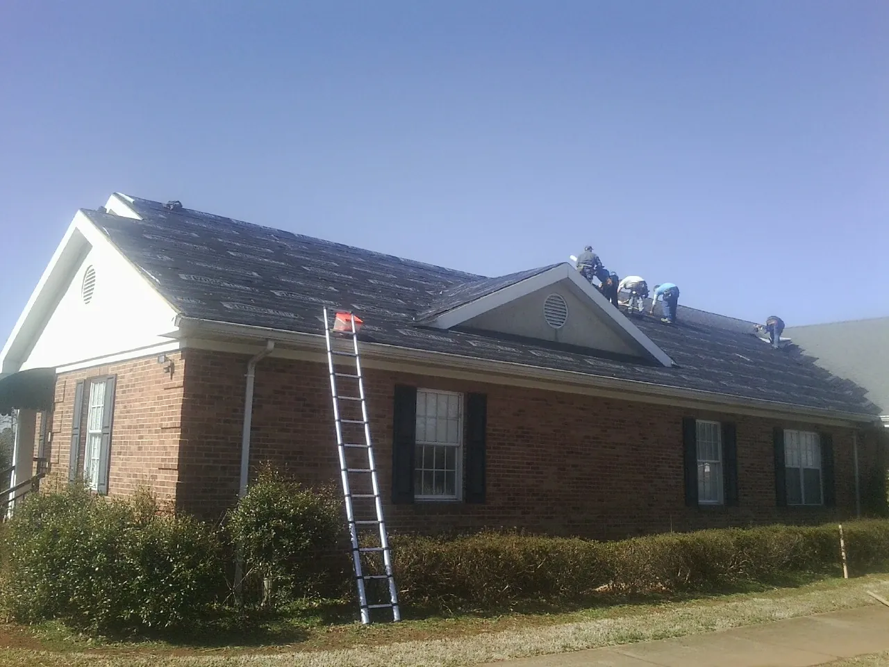 Crew tearing shingles off a brick ranch roof with a ladder set — Metal Roof Repair work by our crew in Hollandale