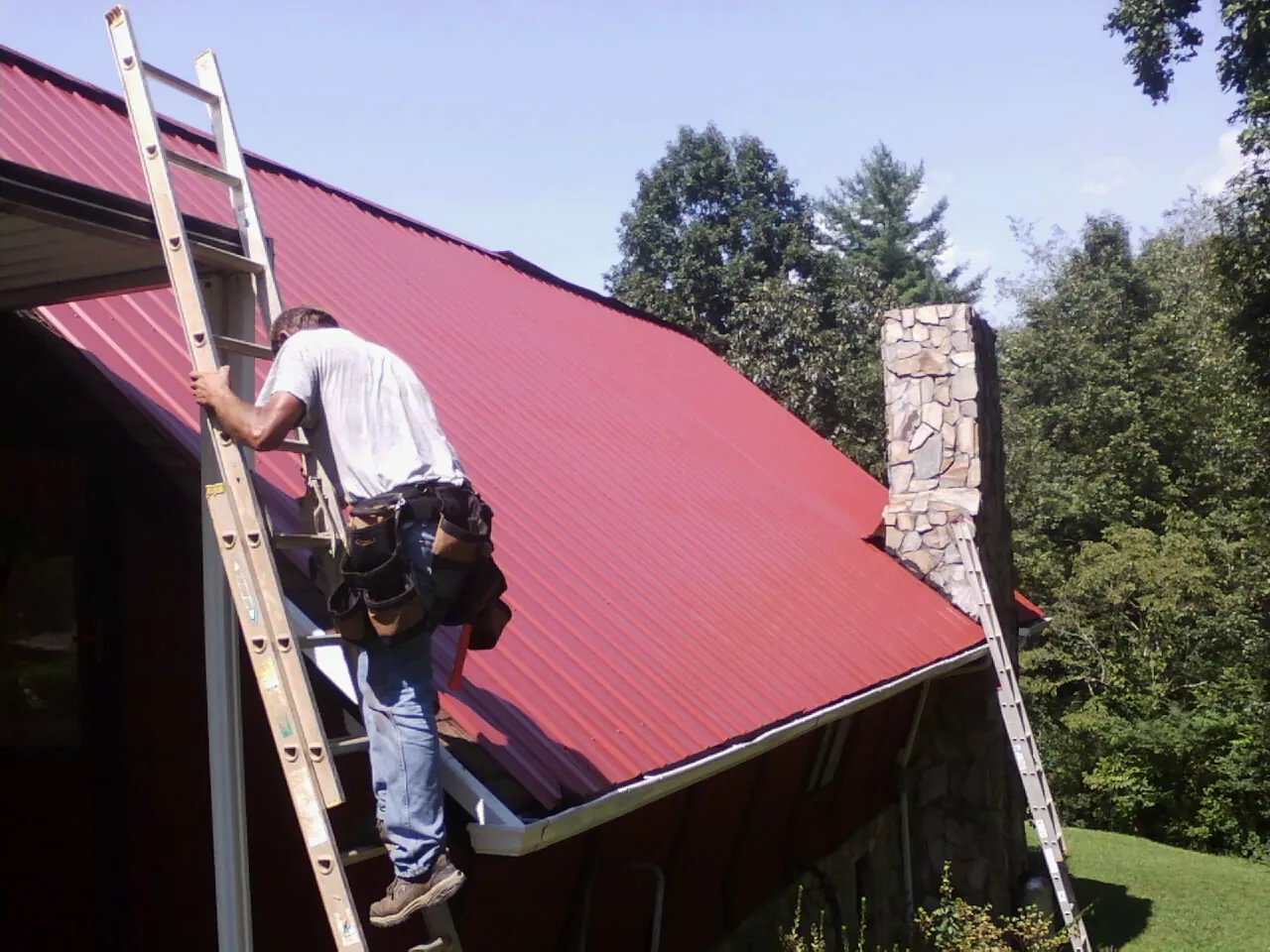 Roofer on a ladder beside a red metal roof and stone chimney — Soffit & Fascia Repair by Roofing Brothers in Hollandale